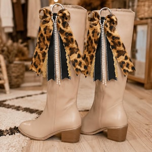 May include: A pair of knee-high beige boots with block heels, featuring decorative attachments at the top. Each boot is adorned with a silver ring holding a leopard print ribbon, a solid black ribbon, a gold lace trim, and strands of white pearls.