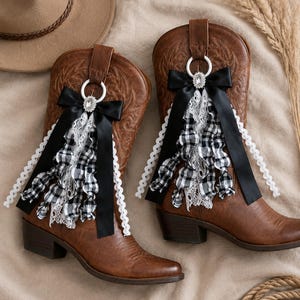 Könnte beinhalten: Ein Paar braune Cowboystiefel aus Leder mit dekorativen Anhängern an den Zuglaschen. Jeder Stiefel ist mit einer schwarzen Satinschleife, weißer Spitzenborte und einer schwarz-weiß karierten Stoffrüsche verziert, mittig mit einem weißen Ring und Kristall.