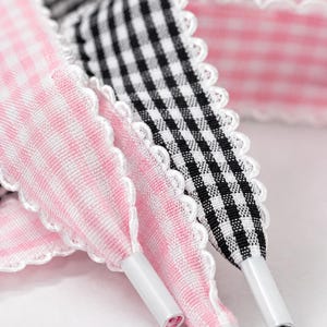 May include: A set of fabric shoelaces featuring a scalloped white edge. One lace has a pink and white gingham pattern, while the other has a black and white gingham pattern. Each lace is finished with a smooth white plastic aglet at the tip.