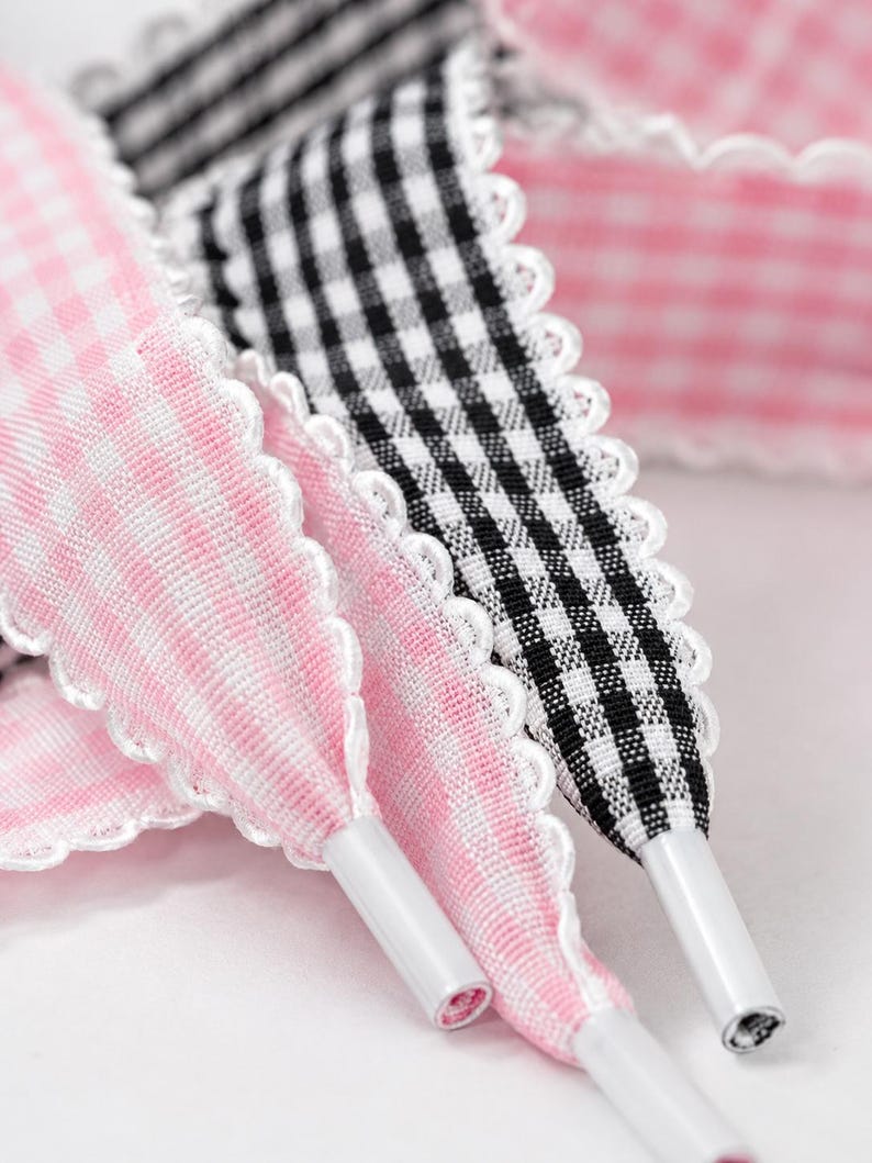 May include: A set of fabric shoelaces featuring a scalloped white edge. One lace has a pink and white gingham pattern, while the other has a black and white gingham pattern. Each lace is finished with a smooth white plastic aglet at the tip.