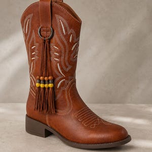 Könnte beinhalten: Ein wadenhoher brauner Stiefel mit weißen Ziernähten am Schaft und an der Schuhspitze. Ein dunkler Metallring ist an der Seite befestigt und hält eine braune Lederquaste, die mit vier Holzperlen in abwechselnd gelben und braunen Farben verziert ist.