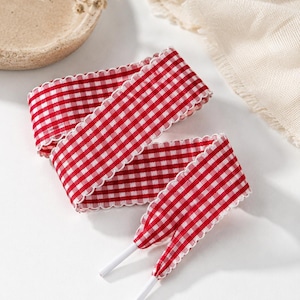 May include: A pair of flat shoelaces featuring a red and white gingham check pattern. The laces have a scalloped edge detail along both sides and are finished with solid white plastic aglets at the ends for easy threading through eyelets.