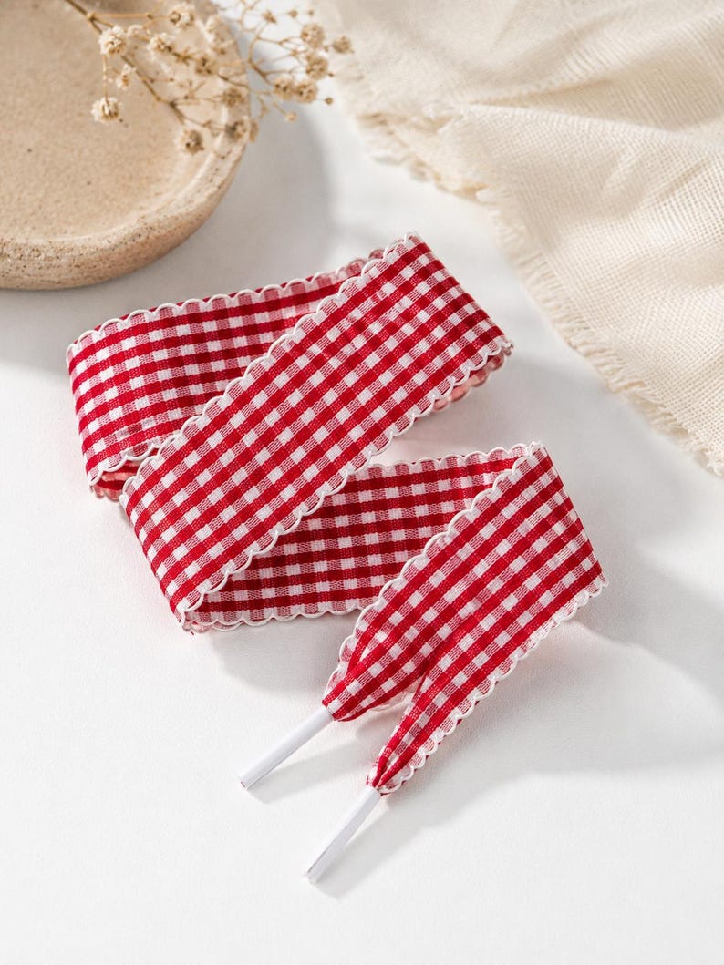 May include: A pair of flat shoelaces featuring a red and white gingham check pattern. The laces have a scalloped edge detail along both sides and are finished with solid white plastic aglets at the ends for easy threading through eyelets.