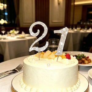 Peut inclure: Un gâteau rond au glaçage blanc garni de dés de pêches, d'une fraise et de petites perles blanches décoratives. Une décoration de gâteau argentée en forme de chiffre 21, incrustée de strass transparents, est plantée sur le dessus du gâteau.