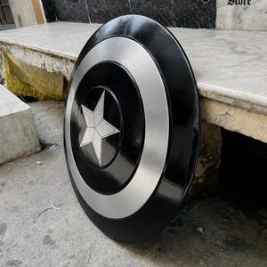 May include: A black and silver shield with a white star in the center. The shield has a circular design with a silver ring around the star and a black outer edge. The shield is leaning against a stone surface.