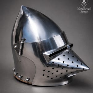 May include: A polished steel houndskull bascinet helmet featuring a pointed visor with a horizontal eye slit and numerous ventilation holes. The side displays a metal hinge mechanism. The text Medieval Store is visible in the upper right corner.
