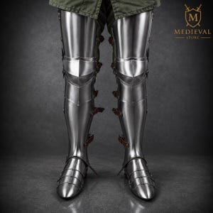 May include: Polished steel leg armor featuring articulated cuisses, poleyns, greaves, and sabatons. The armor is secured with brown leather straps and buckles along the sides. The logo for Medieval Store is visible in the upper right corner.