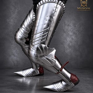 May include: Polished steel leg armor featuring articulated thigh plates, knee cops, and greaves with pointed sabatons. The metal pieces are secured with rivets and brown leather straps. The logo for Medieval Store is visible in the top right corner.