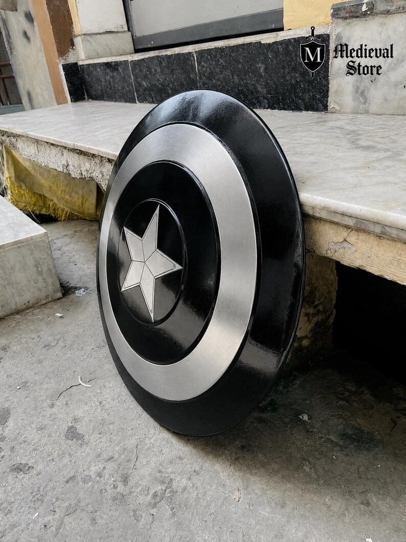 May include: A black and silver shield with a white star in the center. The shield has a circular design with a silver ring around the star and a black outer edge. The shield is leaning against a stone surface.