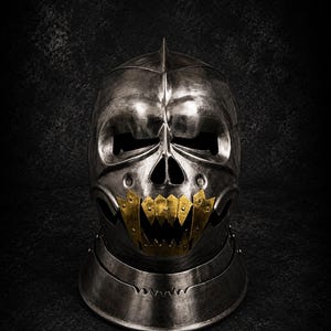 Könnte beinhalten: Ein dunkler Stahlhelm mit einer schädelartigen Frontplatte, einem Mittelgrat, horizontalen Augenschlitzen und einer Nasenöffnung. Der Unterkieferbereich ist mit gezackten, goldfarbenen Metallzähnen verziert. Der Text MEDIEVAL STORE steht oben rechts.