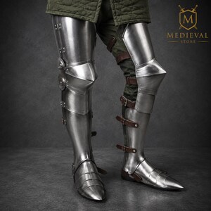 May include: Polished steel leg armor featuring articulated greaves, poleyns with side discs, and sabatons. The metal plates are secured with brown leather straps and buckles, covering the lower limbs from the thigh down to the pointed feet.