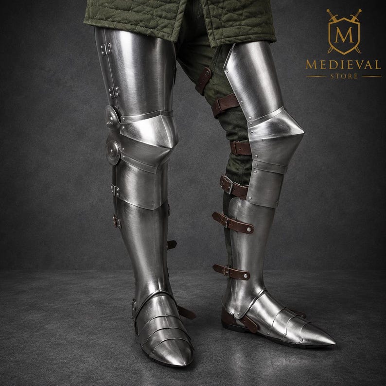 May include: Polished steel leg armor featuring articulated greaves, poleyns with side discs, and sabatons. The metal plates are secured with brown leather straps and buckles, covering the lower limbs from the thigh down to the pointed feet.