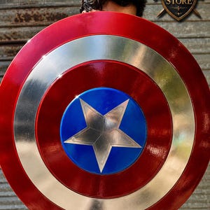 Pu&ograve; includere: Uno scudo rosso, argento e blu con un'emblema a stella. Lo scudo ha un design circolare con un anello esterno rosso, un anello centrale argentato e un centro blu con una stella argentata. Lo scudo &egrave; tenuto davanti a una persona.