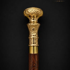 May include: A walking stick featuring a polished gold-tone metal handle with intricate floral engravings and a rounded top. The handle sits atop a dark brown wooden shaft with a visible grain pattern. The text MEDIEVAL STORE appears in the corner.