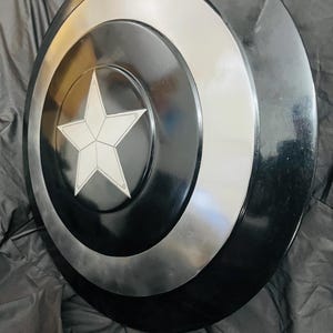May include: A round, black and silver shield with a white star in the center. The shield has concentric circles in black and silver. The star is outlined in white. The shield is a prop or collectible.