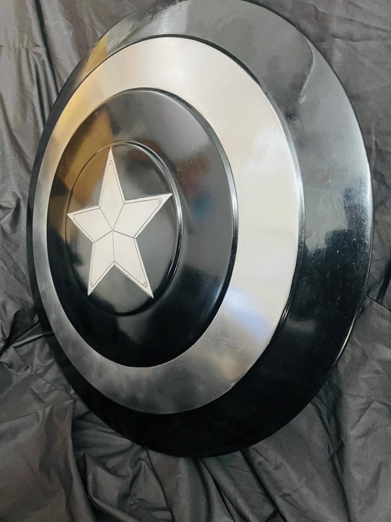 May include: A round, black and silver shield with a white star in the center. The shield has concentric circles in black and silver. The star is outlined in white. The shield is a prop or collectible.