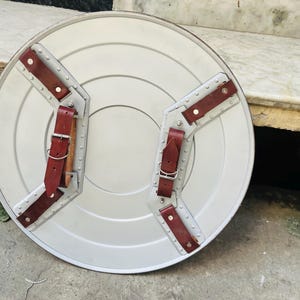 May include: A silver-colored, round shield with a central design and riveted metal supports. It features two brown leather straps with buckles for securing the shield. The shield is likely a prop or decorative item.