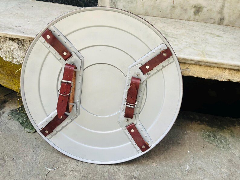 May include: A silver-colored, round shield with a central design and riveted metal supports. It features two brown leather straps with buckles for securing the shield. The shield is likely a prop or decorative item.