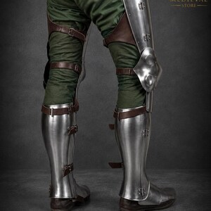 May include: Polished steel leg armor featuring articulated knee plates and greaves secured with brown leather straps and metal buckles over green fabric trousers. The armor includes a logo with the text MEDIEVAL STORE in the upper right corner.