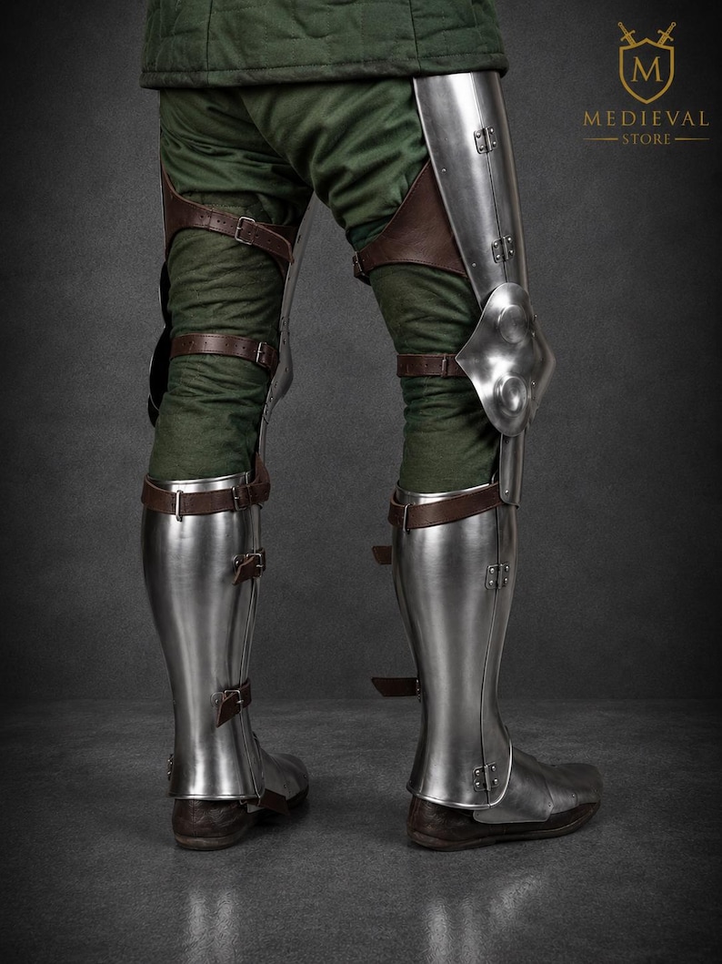May include: Polished steel leg armor featuring articulated knee plates and greaves secured with brown leather straps and metal buckles over green fabric trousers. The armor includes a logo with the text MEDIEVAL STORE in the upper right corner.