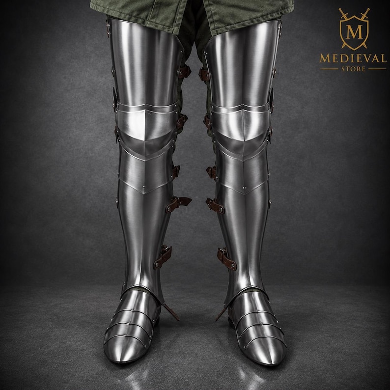 May include: Polished steel leg armor featuring articulated cuisses, poleyns, greaves, and sabatons. The armor is secured with brown leather straps and buckles along the sides. The logo for Medieval Store is visible in the upper right corner.