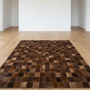 Puede incluir: Una alfombra rectangular grande con un diseño de patchwork. La alfombra está hecha de cuadrados en varios tonos de marrón, creando un patrón de tablero de ajedrez. La alfombra está colocada sobre un suelo de madera en una habitación con paredes blancas.