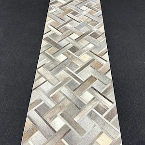 May include: A rectangular runner rug featuring a geometric basketweave pattern composed of individual strips of cowhide in varying shades of beige, tan, gray, brown, and white, creating a textured surface with a multi-tonal appearance.