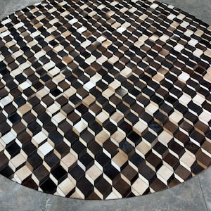 May include: A large circular rug composed of small, individual square tiles of cowhide leather. The tiles are arranged in a repeating geometric pattern of black, white, and various shades of brown, creating a three-dimensional cube effect.