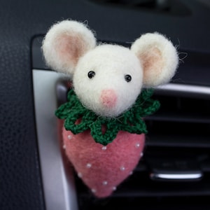 May include: A needle-felted white mouse figure with black eyes and a pink nose, wearing a dark green crocheted collar. The mouse is attached to a pink, strawberry-shaped body decorated with small white beads, designed as a car air vent clip.