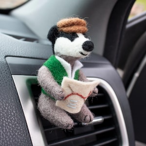 May include: A needle-felted badger figurine wearing a brown cap and a green vest, holding a small beige map with red and blue stitched details. The badger has black and white facial markings and grey fur, designed to attach to a car air vent.