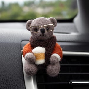 May include: A grey felt teddy bear wearing gold-rimmed glasses, an orange sweater, and a brown scarf, holding a coffee cup. The bear is designed to clip onto a car vent, adding a touch of whimsy to the vehicle's interior.
