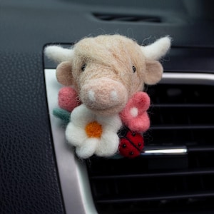 May include: A needle-felted Highland cow car air freshener clip featuring a beige fuzzy cow head with white horns, adorned with a cluster of pink and white felt flowers with orange centres and a small red ladybug with black spots.