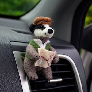 May include: A felted badger figurine wearing a brown cap, green vest, and holding a map. The badger is attached to a car vent, with a black and silver dashboard in the background. The badger has a black and white face and a friendly expression.