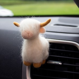 May include: A needle-felted white Highland cow car air freshener clip with a fuzzy texture, featuring light brown horns and matching brown hooves. The small, fluffy animal figure is attached to a black plastic car air vent slat.