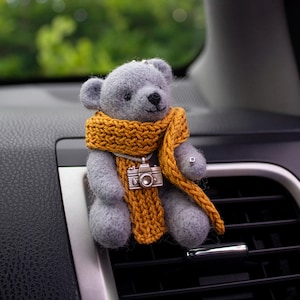 May include: A gray teddy bear car air vent clip with a mustard yellow scarf and a silver camera pendant. The bear is made of soft material and has a black nose and eyes. The scarf is knitted, and the camera pendant adds a unique touch.