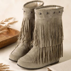 May include: A pair of mid-calf boots in a muted olive green suede finish. The boots feature two distinct layers of long, vertical fringe detailing around the shaft and a row of small, dark metallic studs encircling the top collar of each boot.