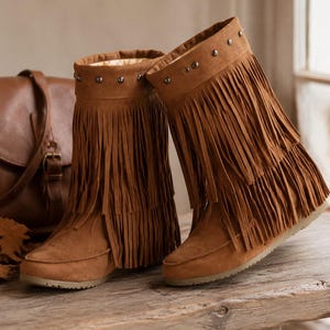 May include: A pair of tan suede mid-calf boots featuring two tiers of long fringe detailing around the shaft. The top of each boot is accented with a row of small, round, metallic studs. The boots have a rounded toe and a flat, textured sole.