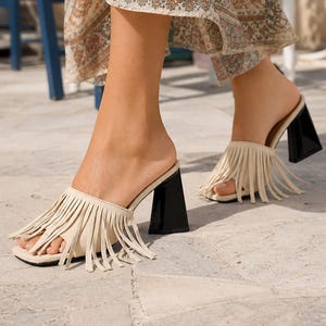 May include: A pair of cream-colored open-toe mules featuring long fringe detailing across the top strap and a solid black, flared block heel. The footwear has a square toe design and a slip-on style, providing a distinct contrast between the light fringe and the dark, structured heel.