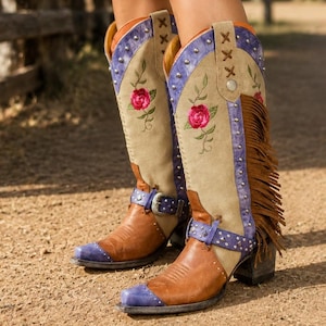 May include: Knee-high boots featuring a tan suede shaft with embroidered pink roses, purple leather accents, silver-tone studs, and brown leather fringe. The boots include a brown leather foot, a purple toe cap, and a buckled strap across the ankle.