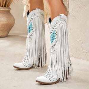 May include: White leather cowboy boots featuring long fringe detailing, silver-toned metal studs, and turquoise blue geometric patterned inlays. The boots have a pointed toe, a stacked wooden heel, and pull tabs at the top of the shaft.