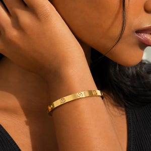 Könnte beinhalten: Ein goldfarbener Metallarmreif mit einer glatten, polierten Oberfläche. Das Band ist mit einem sich wiederholenden Muster aus kleinen, gravierten vierblättrigen Blumenmotiven verziert, die sich mit runden, klaren, funkelnden Steinen abwechseln.