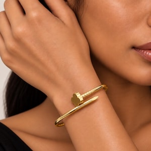 May include: A gold-toned metal bangle bracelet shaped like a bent nail. The head of the nail features a four-leaf clover design. The bracelet has a smooth, polished finish and wraps around the wrist with the pointed end overlapping the clover head.