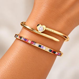 May include: Two gold-toned bracelets. One is a nail-shaped bangle featuring a heart-shaped setting with a clear round stone. The second is a slim, rigid bangle inlaid with a continuous row of square-cut multicolored gemstones in red, blue, purple, and white.
