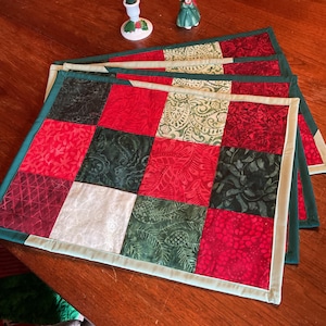 May include: A set of rectangular fabric placemats featuring a patchwork design of square fabric pieces in shades of red, dark green, and cream. The squares display various patterns including paisley, pine needles, and geometric motifs, all bordered by a light green trim.