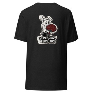 Może przedstawiać: Czarny t-shirt z rysunkiem królika trzymającego bukiet czerwonych róż. Królik ma na sobie bluzę z kapturem i trampki. Pod spodem widnieje napis "EVERYBUNNY NEEDS LOVE".