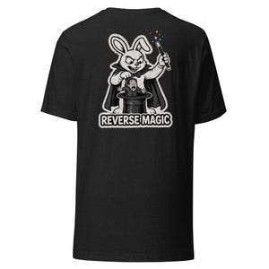 May include: Dark gray t-shirt featuring a black and white illustration of a rabbit magician performing reverse magic. The rabbit is wearing a cape and holding a wand with colorful sparkles. The text "REVERSE MAGIC" is below the illustration.
