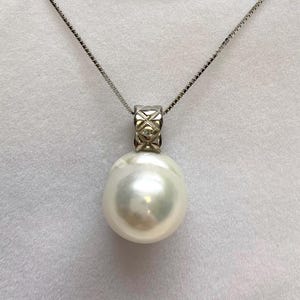 May include: A white spherical pearl pendant suspended from a thin silver chain. The pearl is attached to a metallic bail featuring a textured, cross-hatched pattern. The necklace is displayed on a white velvet-textured card with a small white ribbon.