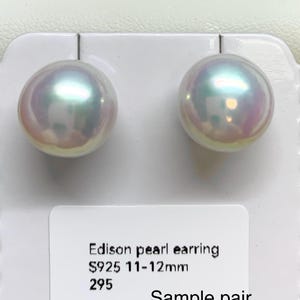 Puede incluir: Un par de pendientes de perlas Edison iridiscentes. Las perlas son redondas, con un diámetro de 1,1 a 1,2 cm. Los pendientes se muestran en una tarjeta blanca. El texto de la tarjeta dice "Edison pearl earring S925 11-12mm Sample pair".