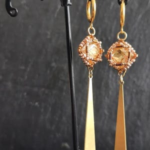 May include: A pair of gold-toned earrings. Each earring features a square, beaded centerpiece with a central amber-colored stone. Below the centerpiece hangs a long, gold-colored, triangular pendant. The earrings are displayed against a dark background.