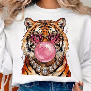 May include: A white sweatshirt featuring a graphic illustration of a tiger wearing heart-shaped glasses and a jeweled necklace while blowing a large pink bubblegum bubble. The tiger has orange and black fur with visible brushstroke textures.